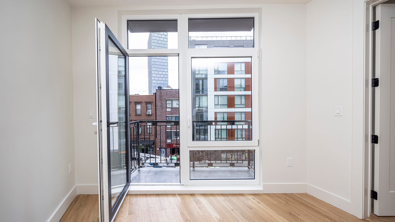 An empty white room with light wood floors and a tall white-framed window wall with an open glass door to a small balcony with a black metal railing, looking across a street at brick buildings with shop signs, a newer white and orange-panelled apartment building and a tall glass tower behind