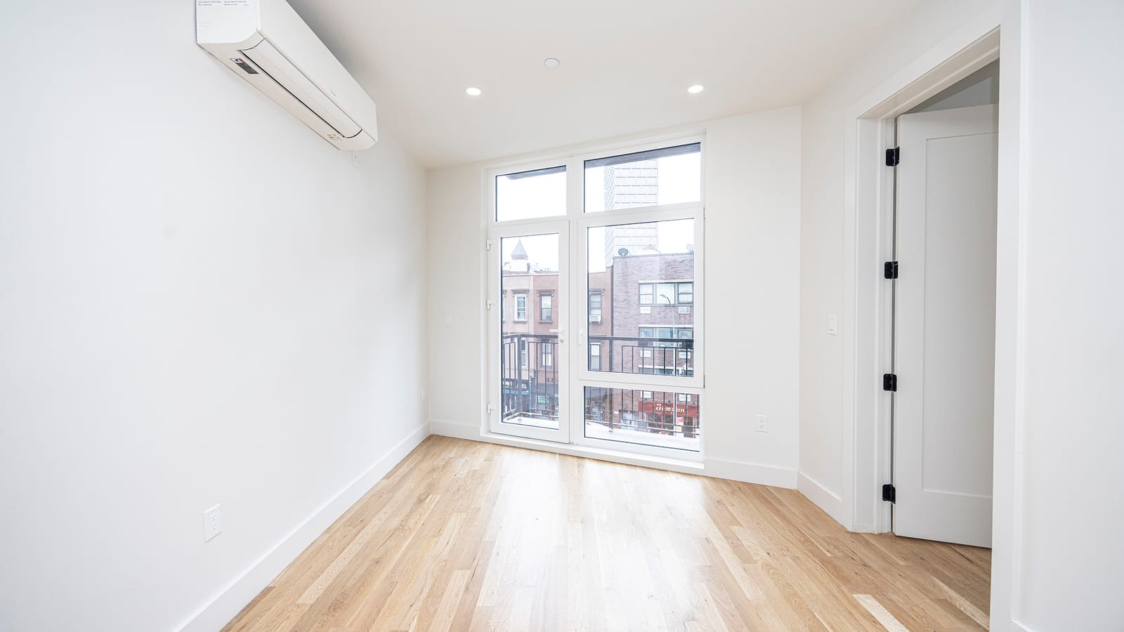 An empty white room with recessed ceiling lights, light wood plank floors, a white wall-mounted heating and cooling unit high on the left wall, an open white door at right and a tall window with a glass door onto a railing, looking out at brick buildings and a tower