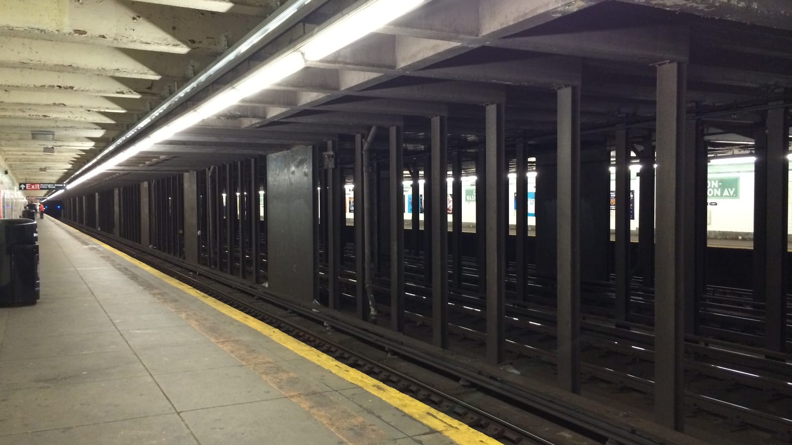 An empty underground subway platform with a yellow edge strip, a row of fluorescent lights and a line of dark steel columns separating several tracks from the far platform, where a green and white sign reads part of the station name