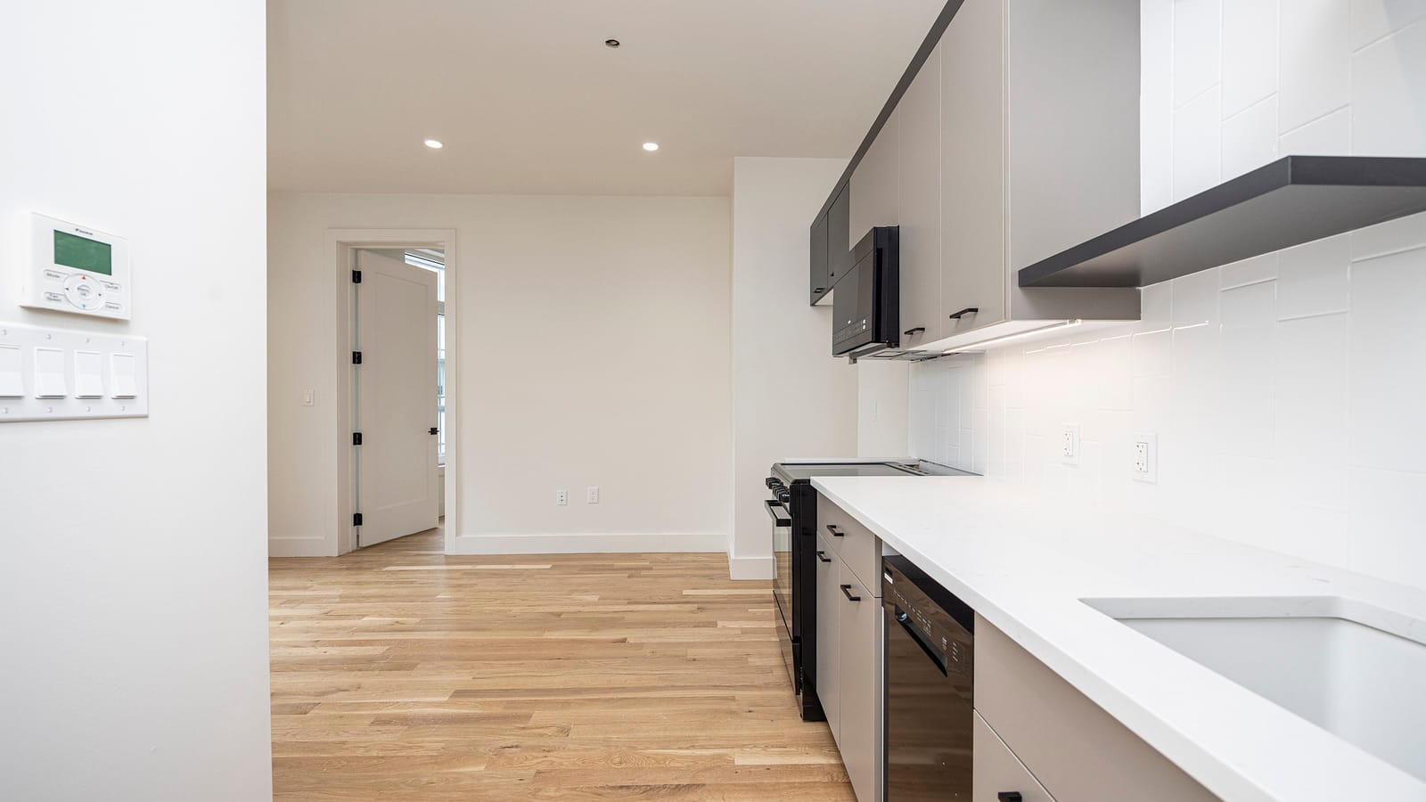A kitchen seen along its counter: a white wall panel with a small screen and buttons and a bank of four light switches at left, light grey cabinets, a dark range hood shelf, a black microwave, a black range with front knobs, a black dishwasher and a white stone counter, with light wood plank floors leading to an empty white room with a doorway