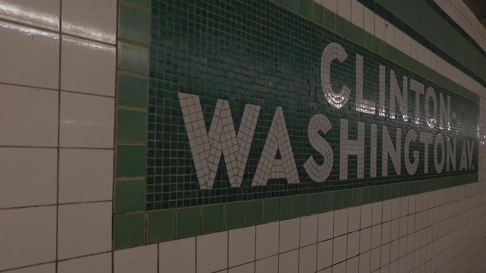 A subway station wall of white tiles with a long dark green mosaic tablet bordered in lighter green, spelling CLINTON-WASHINGTON AV. in small white tiles, seen at an angle