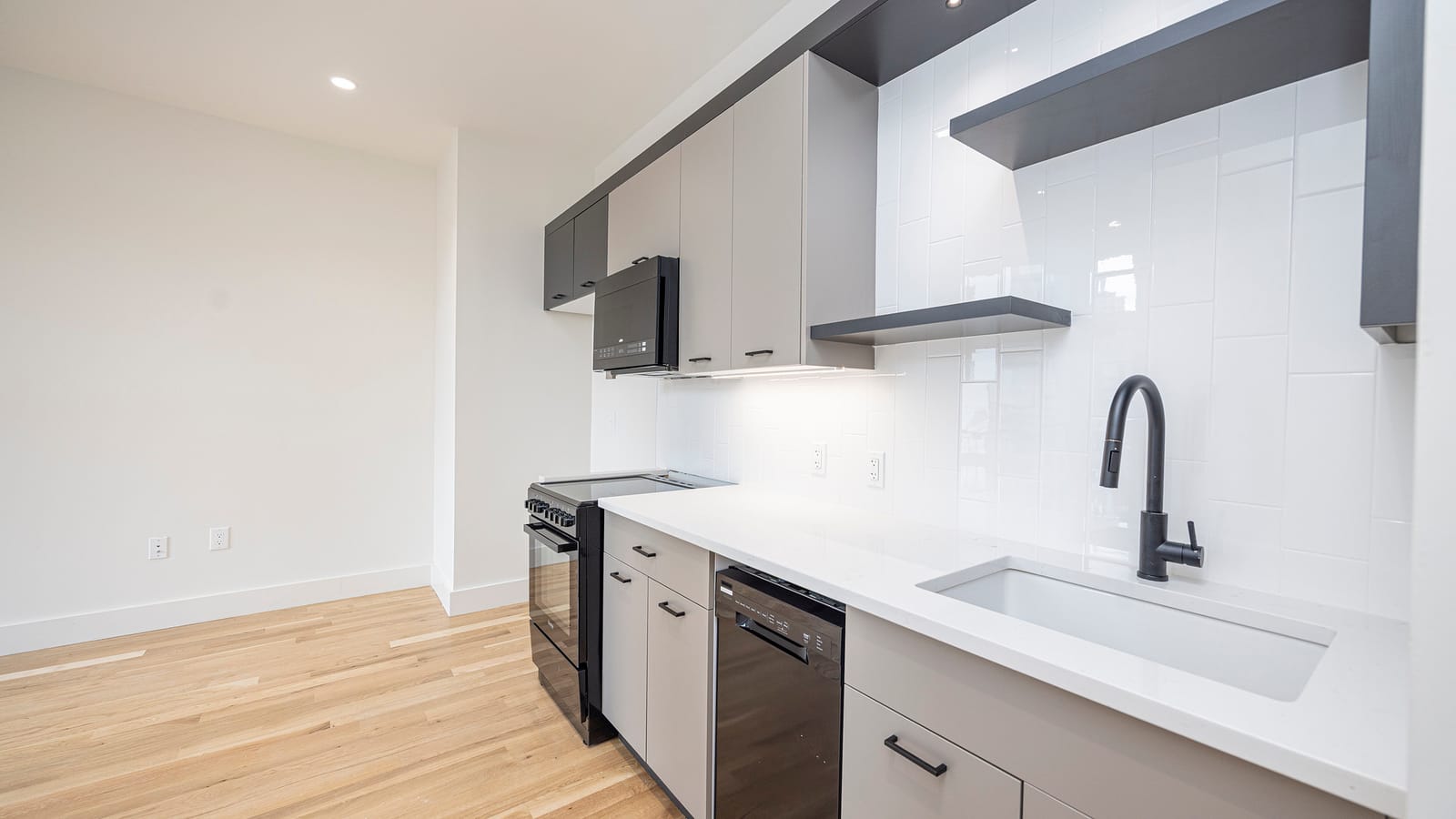 A long kitchen with light grey flat-front cabinets and black handles, a white stone counter with a deep white undermount sink and a black pull-down faucet, white vertical subway tiles, dark floating shelves, a black microwave over a black range with a flat top and front knobs, a black dishwasher, and light wood plank floors running into an empty room