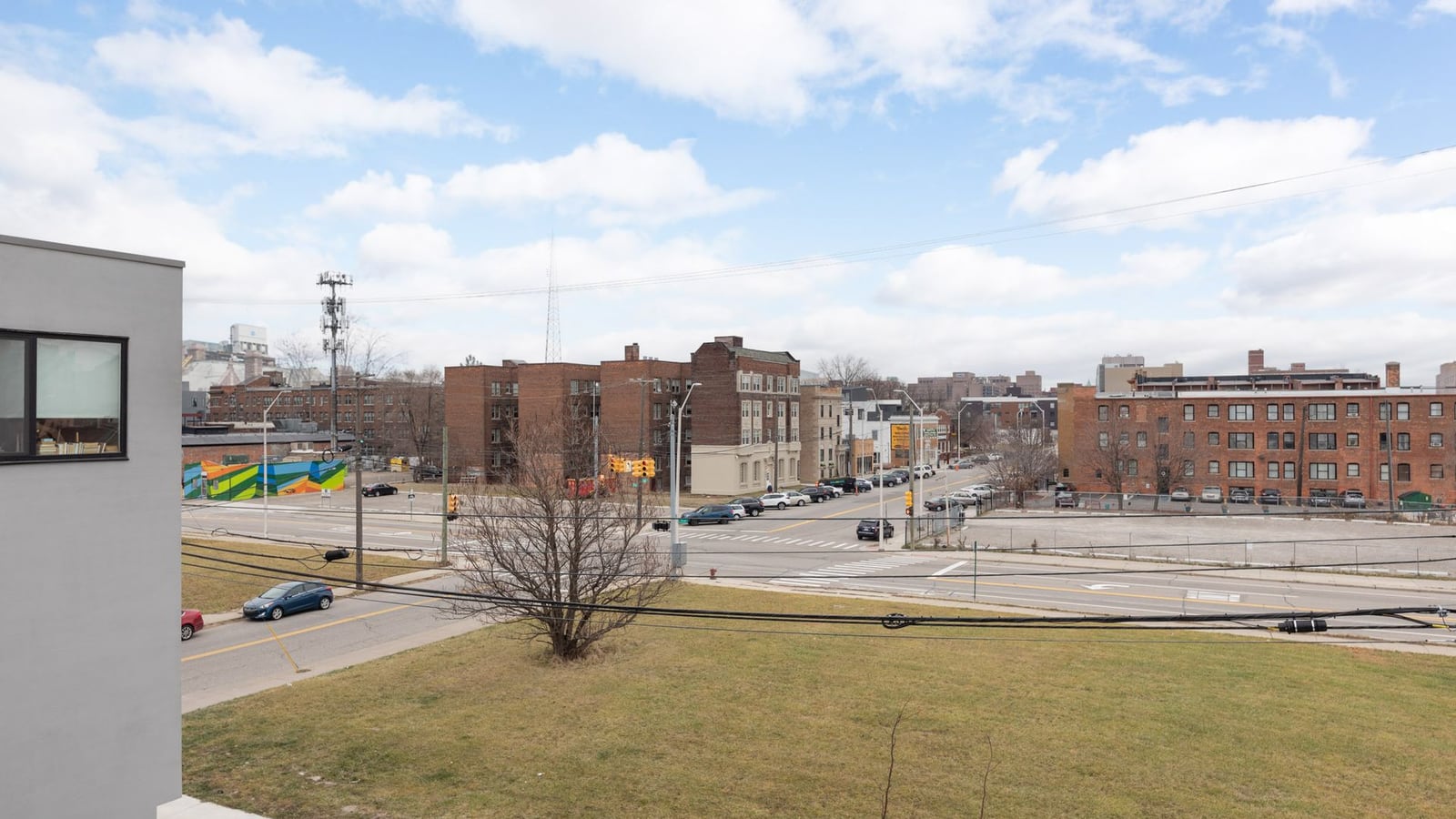 Photograph from an upper floor across a grass lot and a wide street to brick apartment buildings, a painted mural and parked cars under a partly cloudy sky