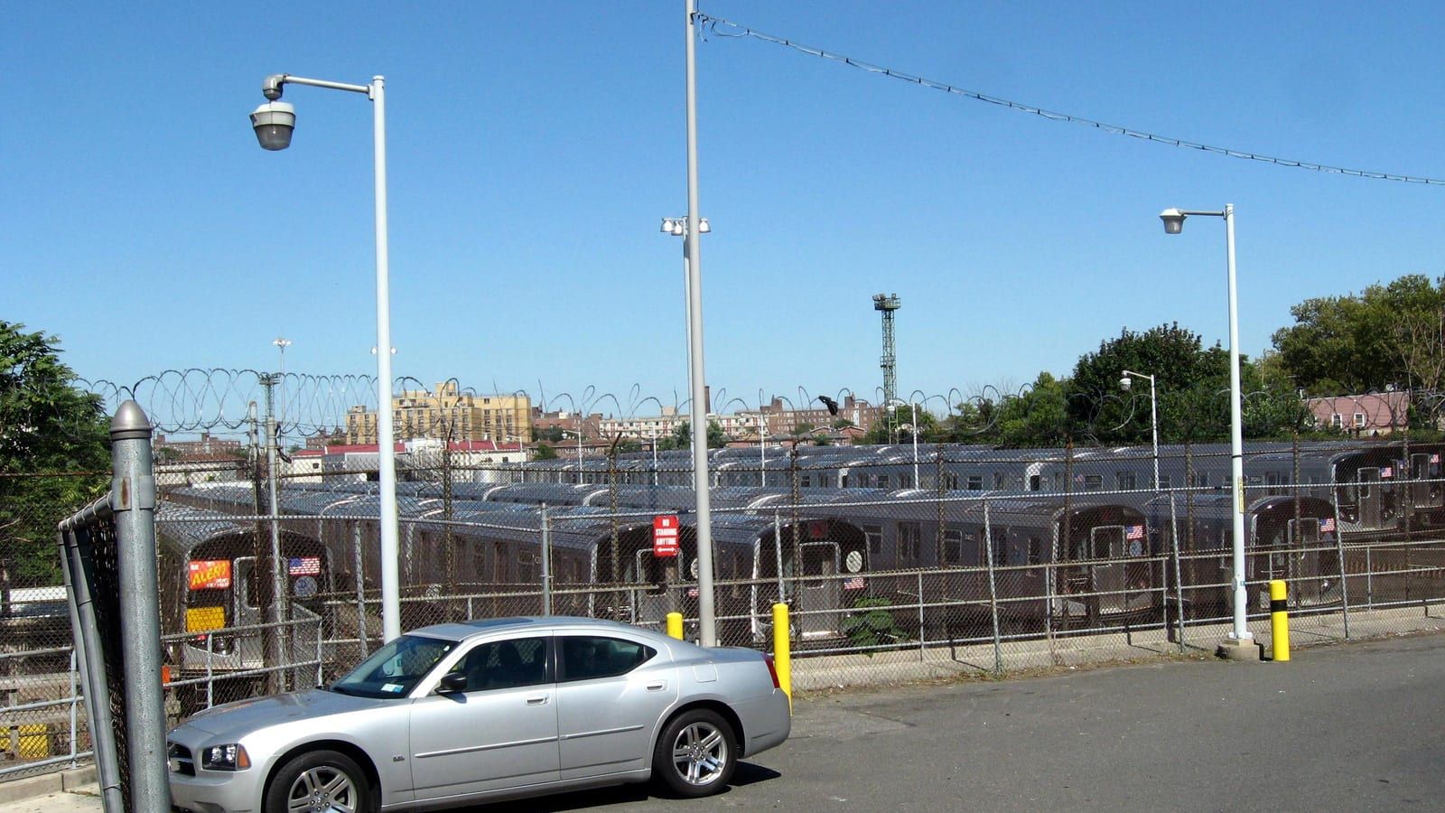Photograph of rows of silver subway cars parked in a rail yard behind a chain-link fence with razor wire, a silver car parked in front, tall light poles, and apartment buildings and trees on the skyline