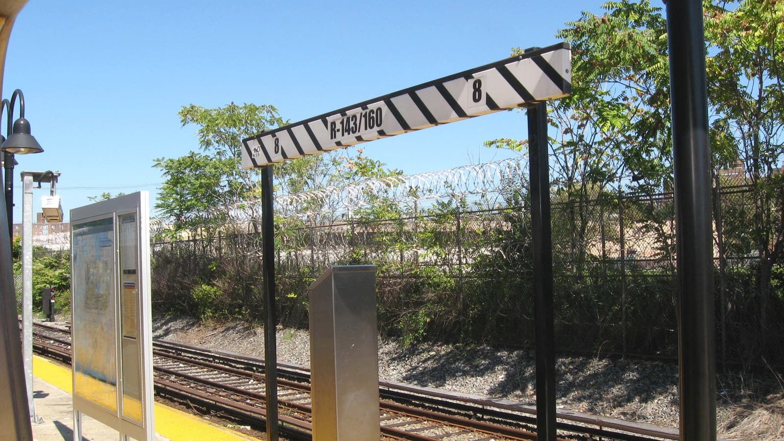 Photograph of an outdoor subway platform with a yellow edge, a map case, a striped car-stop sign reading R-143/160 and 8, tracks below, and a fence topped with razor wire in front of trees under a blue sky