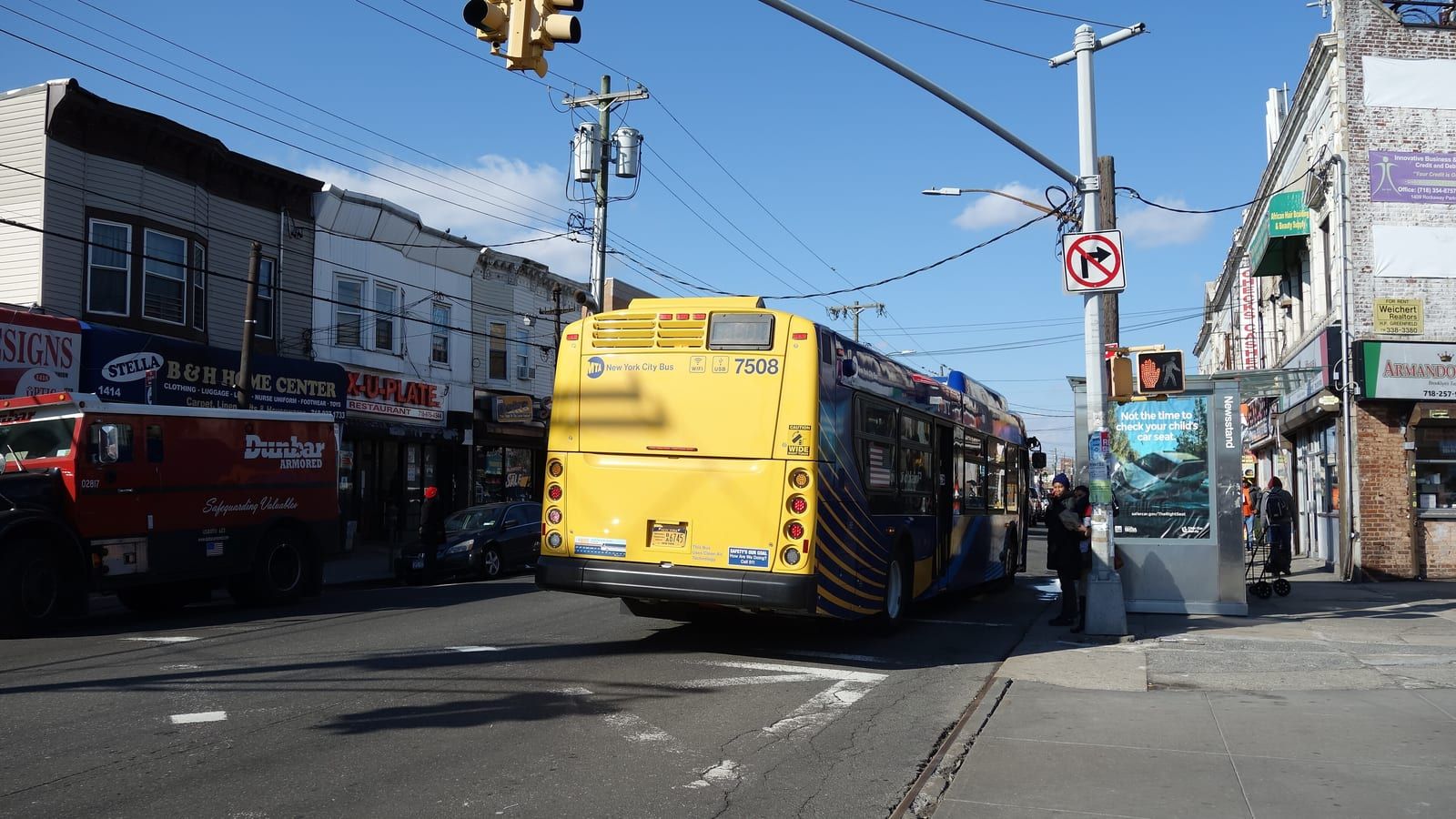 Photograph of a yellow city bus driving away up a two-lane street lined with two-story shop buildings, a red armored truck at left, a bus shelter and a newsstand at right, and traffic lights and wires overhead under a blue sky