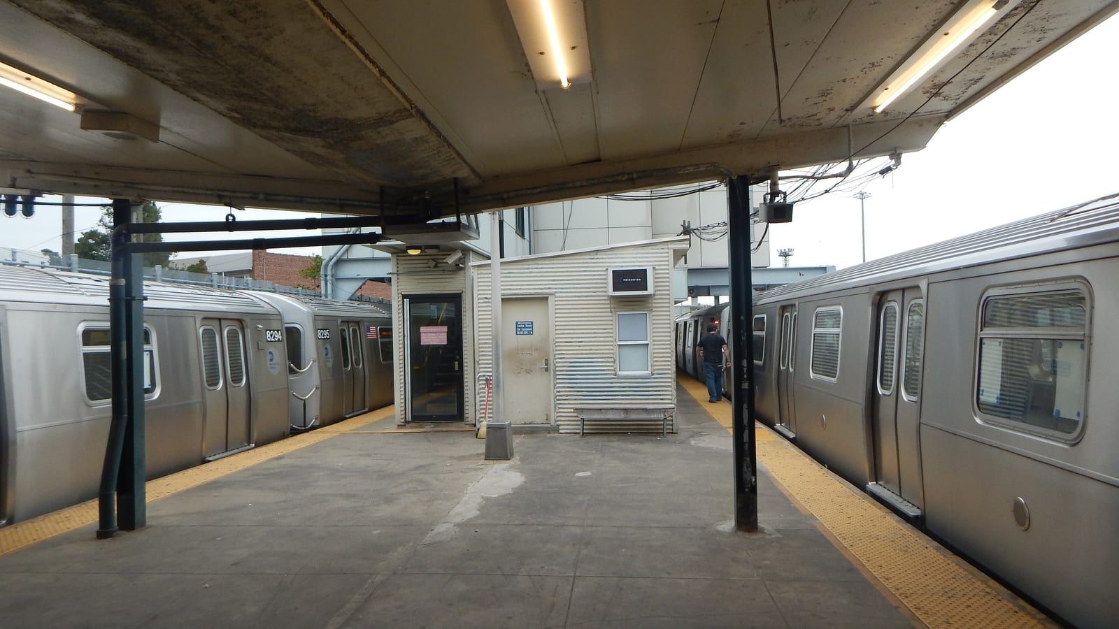 Photograph of a covered subway platform between two silver trains, with a small white corrugated booth and bench in the middle, yellow tactile edges and one person walking away along the right train