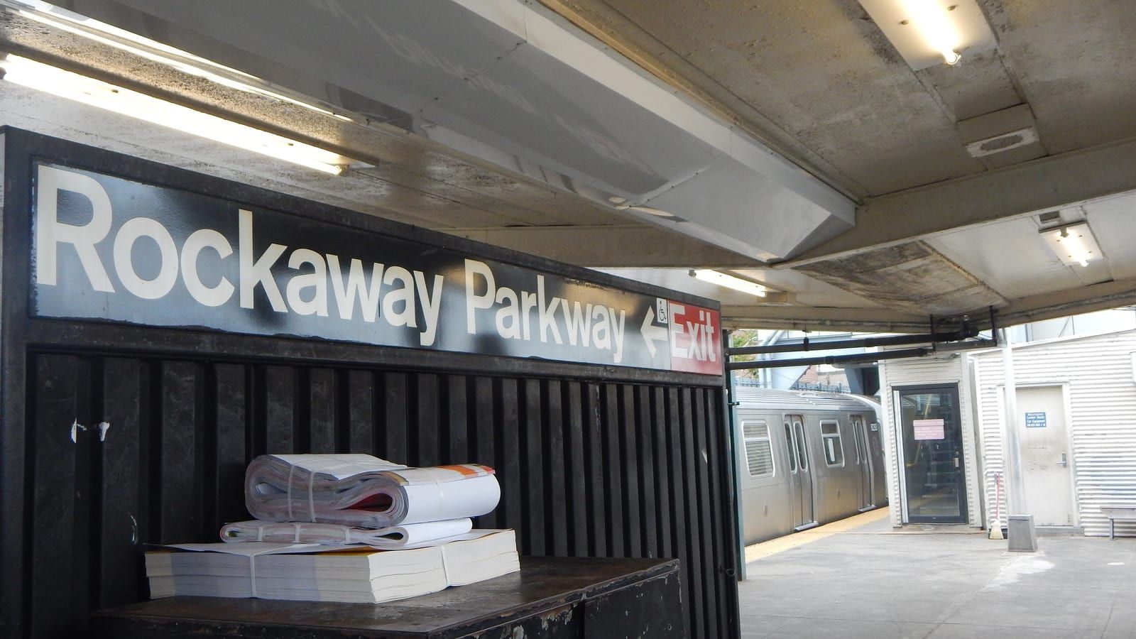 Photograph of a subway platform under a concrete canopy, with a black sign in white letters reading Rockaway Parkway and a red Exit arrow, bundles of papers on a dark metal box, and a silver train and a corrugated white booth in the background