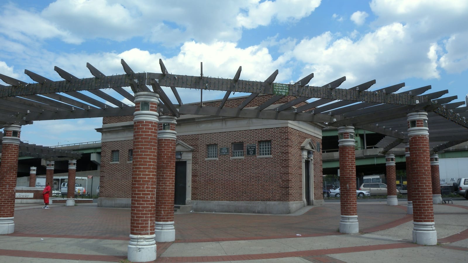 Photograph of a small red brick station house with stone trim in a brick-paved plaza, ringed by brick columns that hold up a wooden pergola, with an elevated highway and parked cars behind it under a blue sky with white clouds