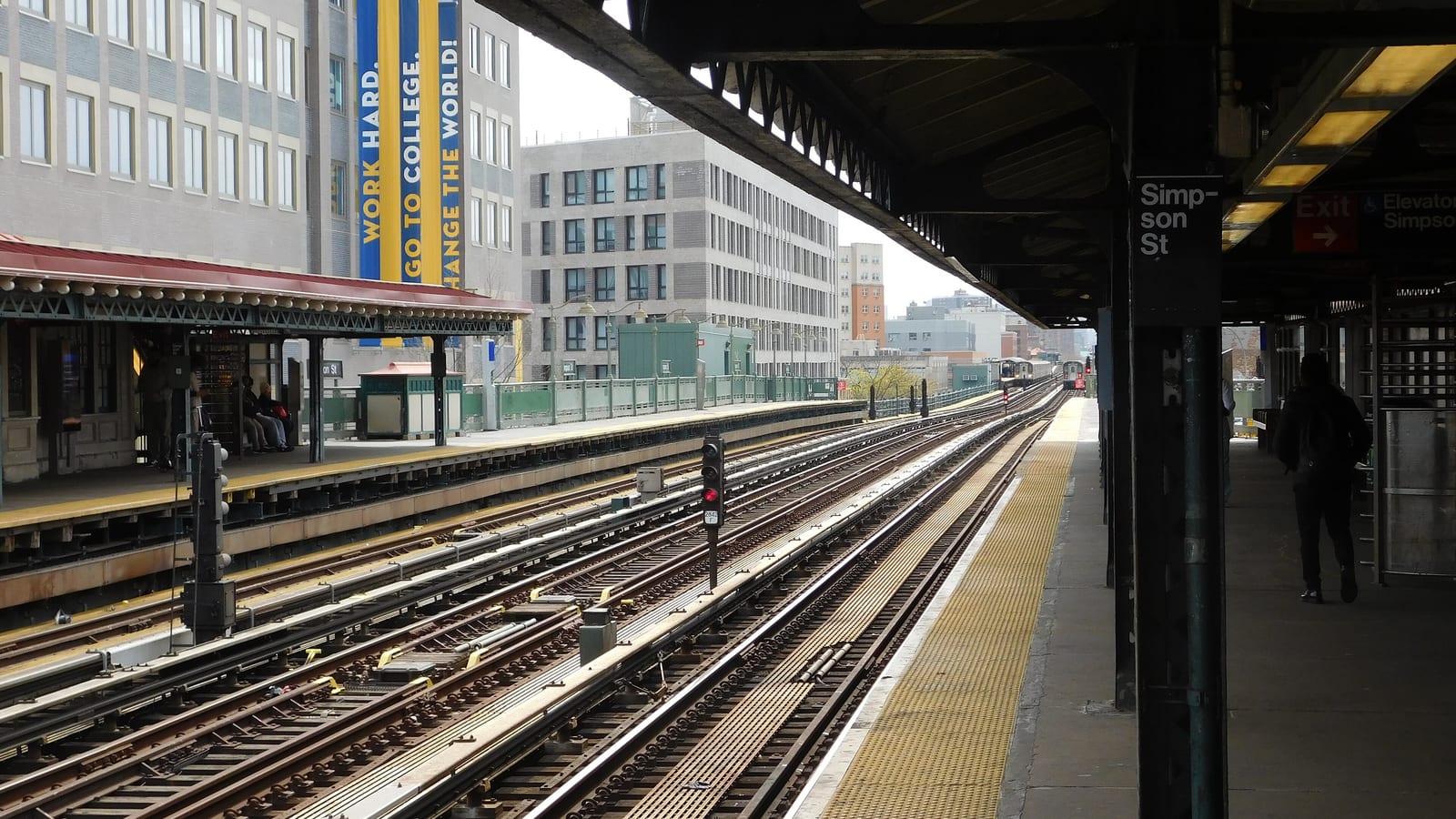 Photograph from an elevated subway platform looking along several tracks, with a sign reading Simpson St on a black column at right, a red-roofed platform canopy at left, and tall pale apartment and school buildings with a large banner behind it