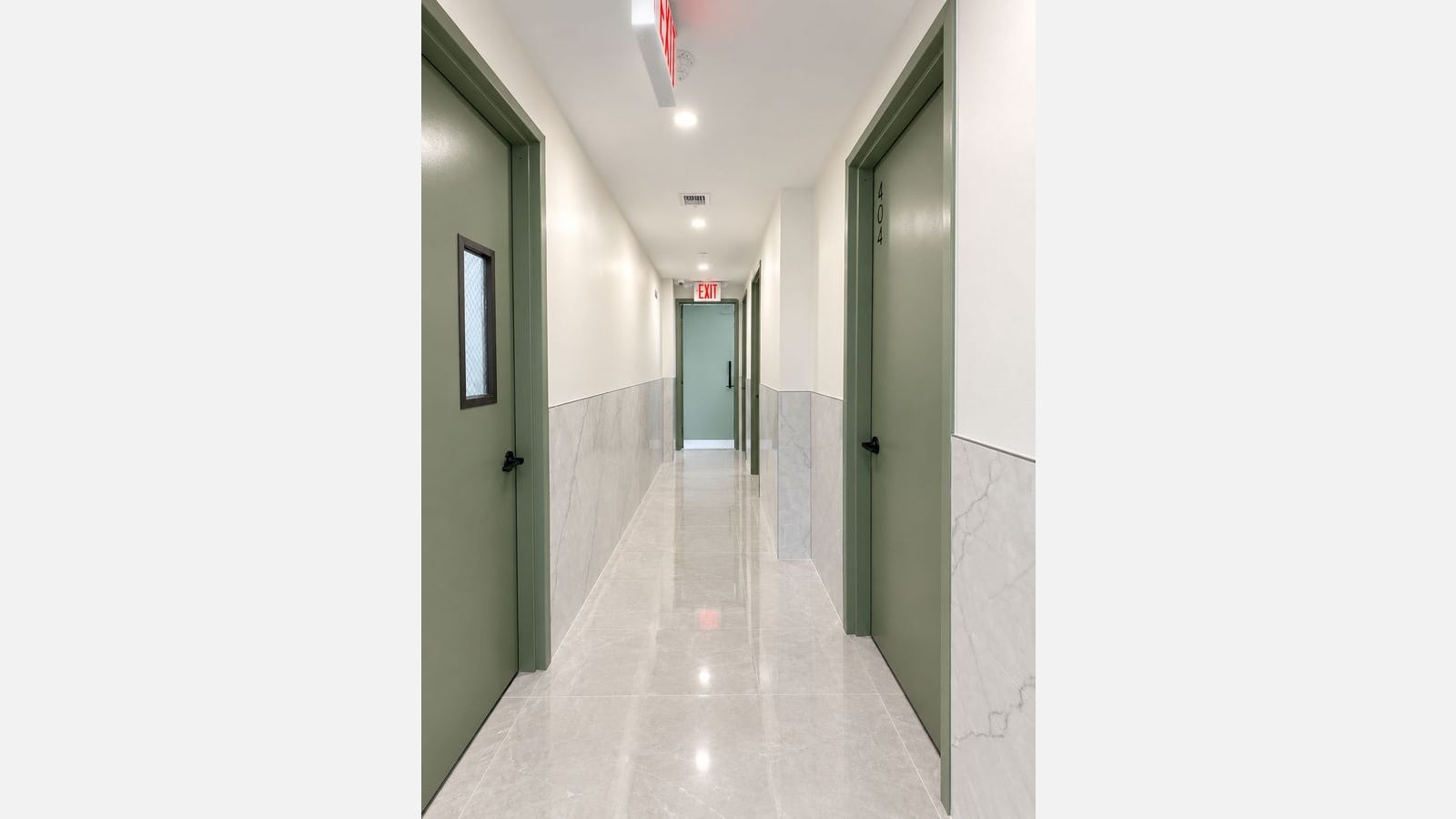 A narrow building hallway with a glossy grey stone-look tile floor, grey marble-look tile on the lower walls and white paint above, green doors and frames on both sides, the right-hand door numbered 404, recessed ceiling lights, and a lit EXIT sign over a green door at the far end