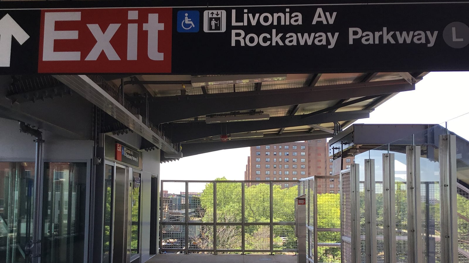 A station platform under a dark overhead sign reading Exit, with a wheelchair symbol, an elevator symbol, Livonia Av, Rockaway Parkway and the L; below it glass elevator doors at left, a fenced platform end with trees and a tall brick apartment building beyond, and glass and steel railings at right