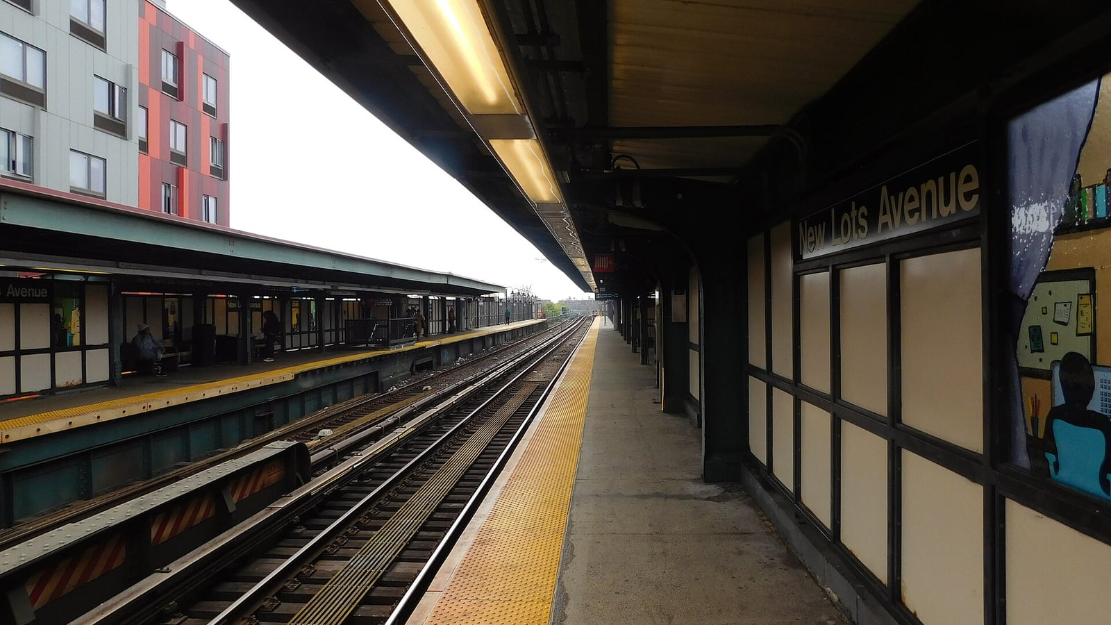 An elevated subway platform under a long canopy with fluorescent lights: a yellow tactile strip runs along the edge beside the tracks, a sign reading New Lots Avenue hangs on the windbreak wall at right beside a painted mural panel, the opposite platform and its shelter are at left, and a grey and red apartment building rises behind it