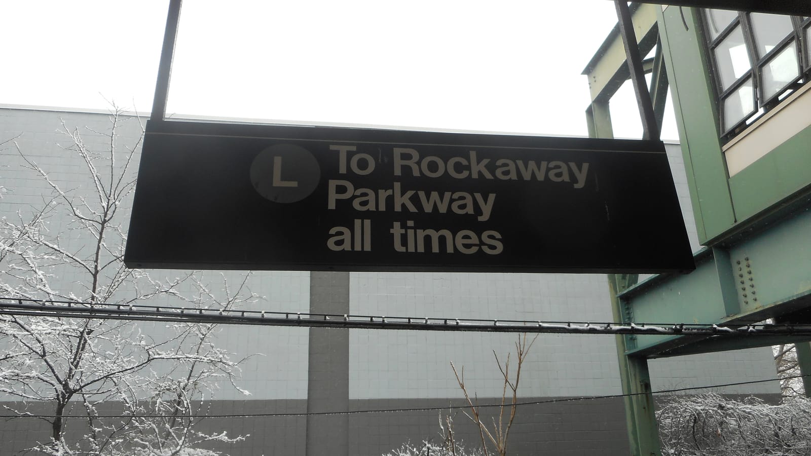 Photograph of a black hanging subway sign with an L symbol reading To Rockaway Parkway all times, with snow on the branches of bare trees, a gray tiled wall behind and the green steel of a station structure at right