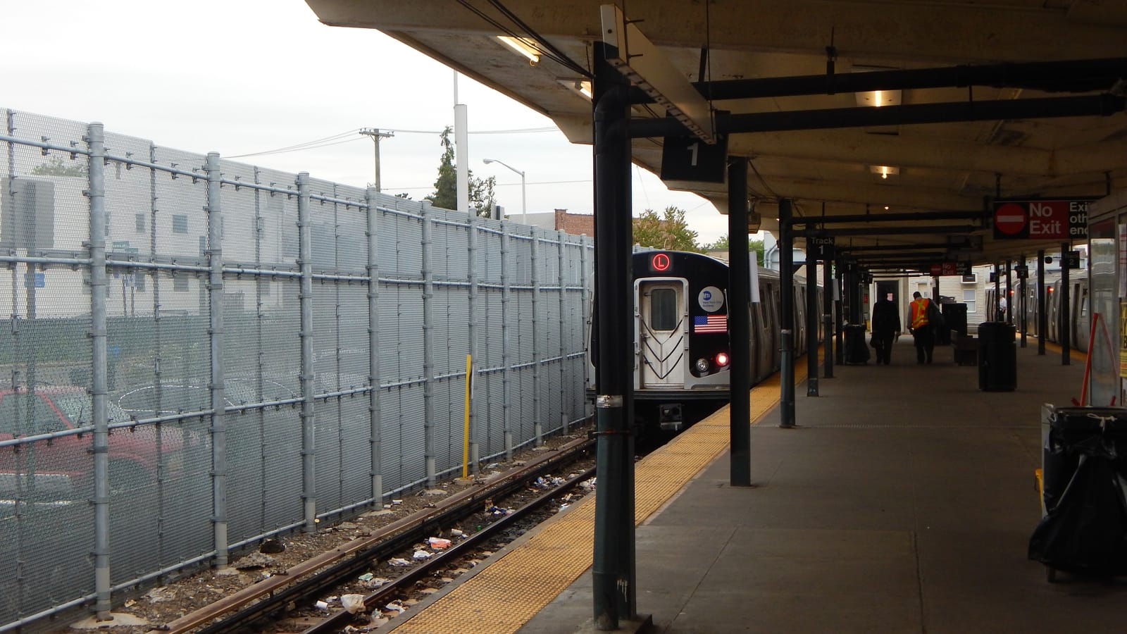 Photograph along a covered outdoor subway platform with a yellow edge strip, an L train with a red L sign and an American flag decal stopped at the platform, a tall metal fence beside the track at left, a Track 1 sign and a red No Exit sign under the canopy, and two people walking away in the distance