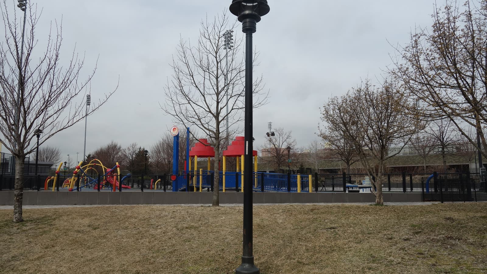 A park lawn of brown winter grass with a black lamp post in front, bare young trees, and behind a black fence a playground with red-roofed towers, blue panels and yellow and red climbing frames, with tall field lights and a gray cloudy sky
