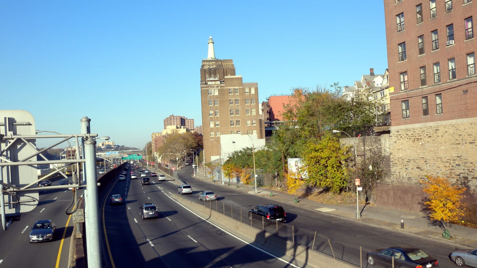 A wide expressway with cars running toward the distance under a clear blue sky, a service road and sidewalk on the right below a stone retaining wall, a tall brown brick building topped by a small white lighthouse-like tower in the middle, a red brick apartment building at right and yellow autumn trees along the road