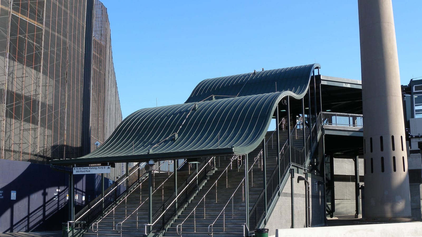 Wide outdoor stairs under a dark green wave-shaped metal canopy leading up to a station bridge, with a sign reading 'To All Trains, Parking, Ferry & Mill Pond Park', a tall concrete column at right and a building wrapped in scaffolding and netting at left, under a clear blue sky