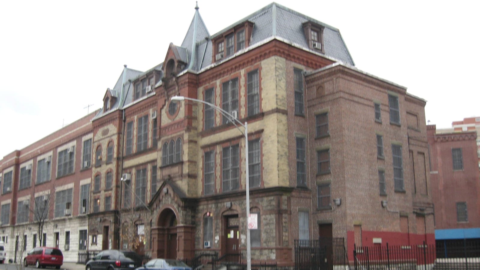 A large old school building of red and tan brick with a gray slate mansard roof, small dormer windows, a pointed corner tower, an arched stone entrance and a black iron fence, with cars parked along the street under a white sky