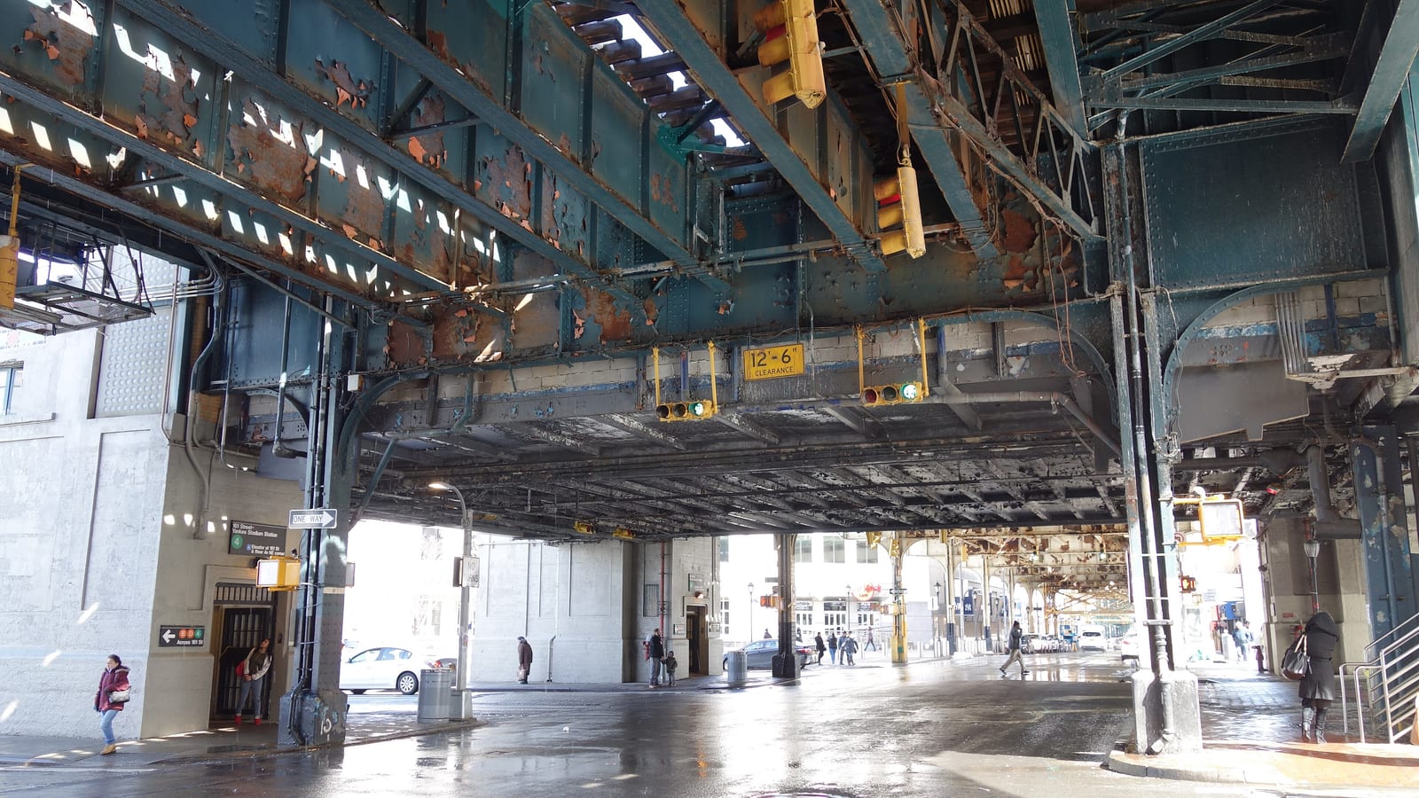 The underside of a raised subway structure with rusted, peeling green steel beams, yellow traffic lights and a yellow '12'-6" clearance' sign over a wet street, a gray station wall at left with a small sign for the 4 train, and a few distant people walking under the tracks