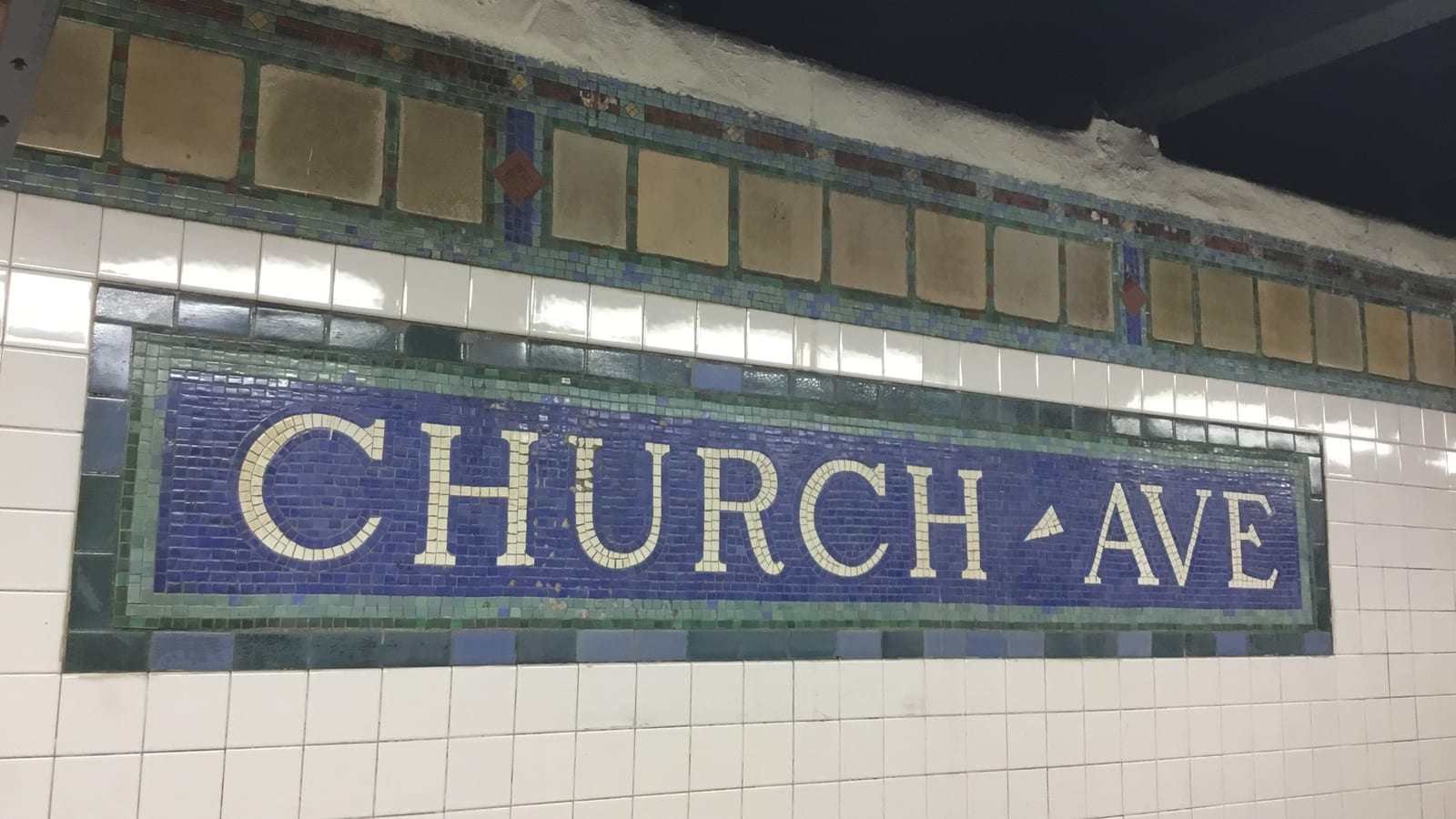 Photograph of a mosaic name tablet on a white tiled subway wall, with cream letters reading CHURCH AVE on a deep blue ground inside a green border, under a band of tan tiles and small colored mosaic squares
