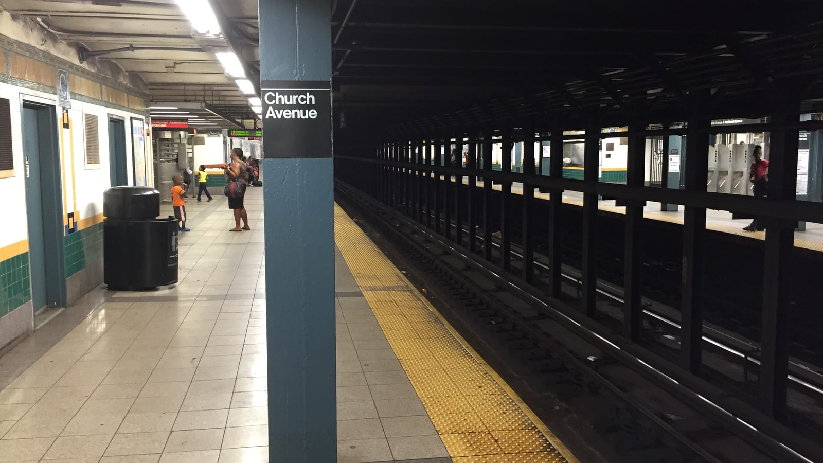 Photograph of an underground subway platform with a blue column carrying a black sign reading Church Avenue, a tiled floor with a yellow edge strip, tracks and black steel columns at right, a trash can and a few small distant figures at left