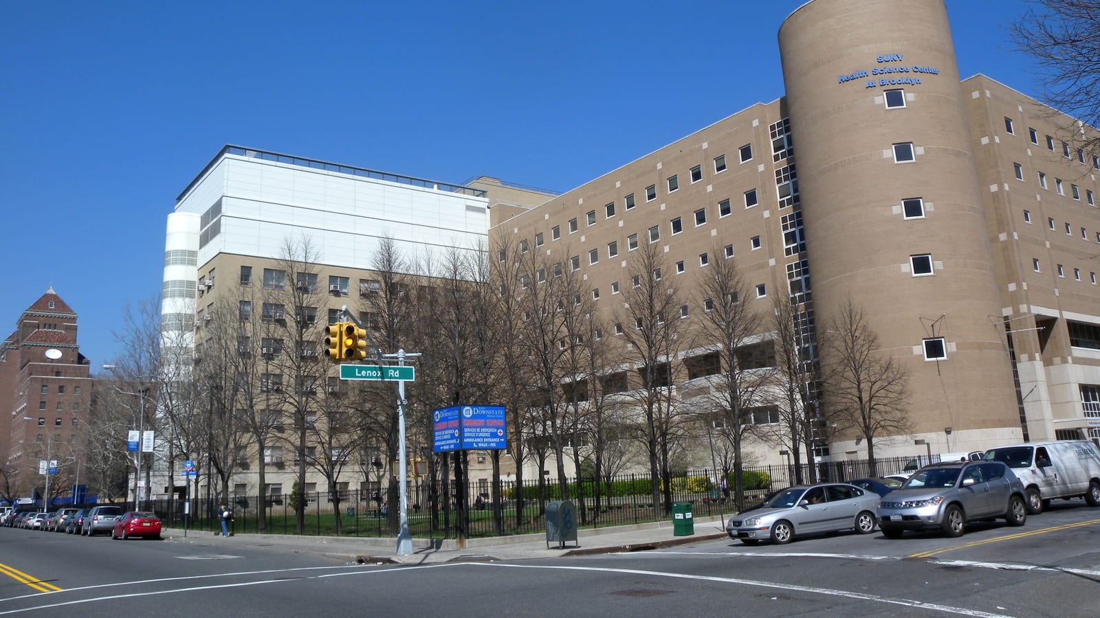 Photograph of a large tan hospital complex with a round tower lettered SUNY Health Science Center at Brooklyn, bare trees behind a black iron fence, a green street sign reading Lenox Rd, blue Downstate emergency signs, parked cars and an empty intersection under a clear blue sky