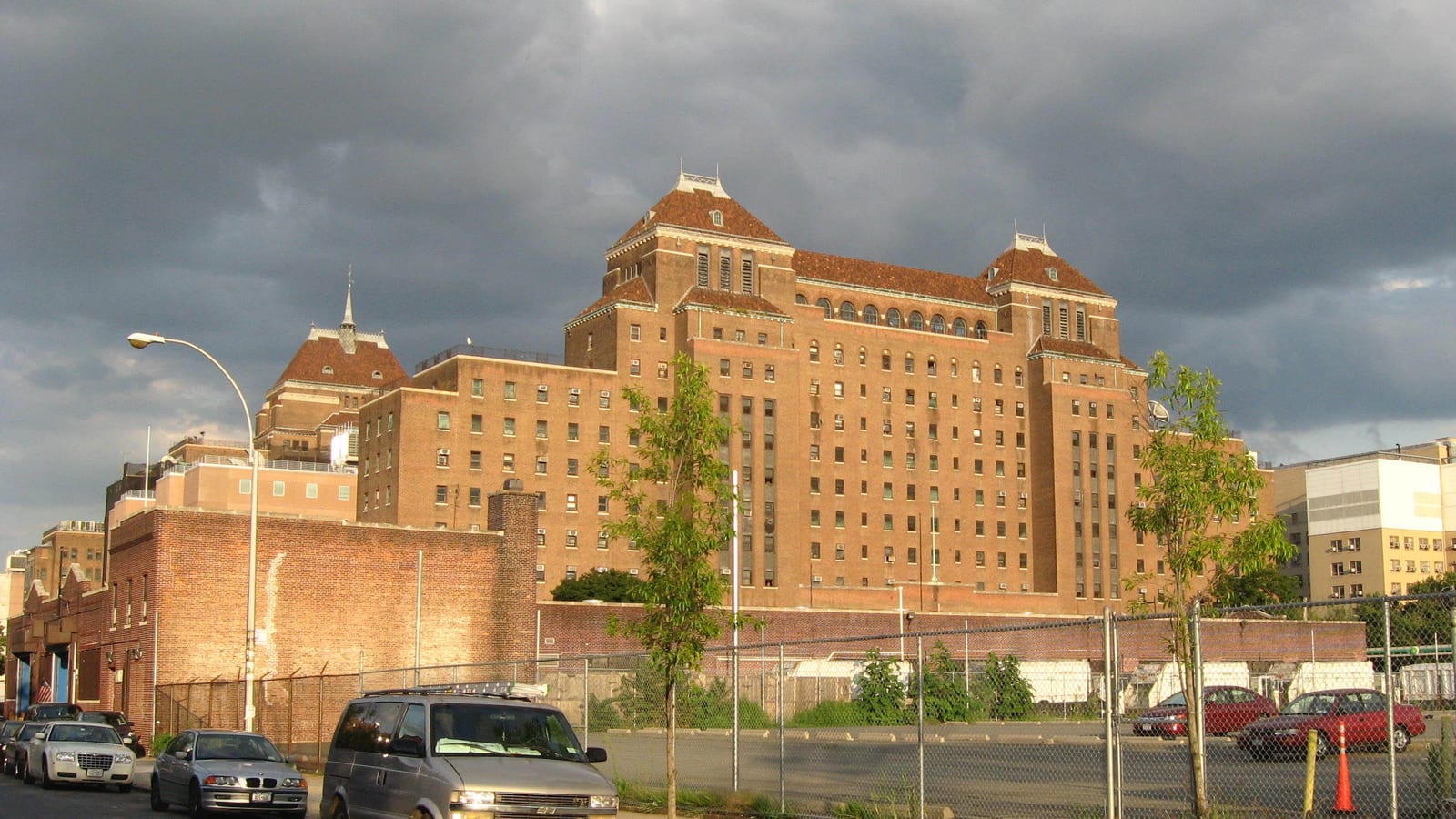 Photograph of a tall brown brick hospital building with peaked tile roofs and towers, lit by low sun under dark storm clouds, behind a brick wall, a chain-link fence, a parking lot and young street trees, with parked cars on the street in front