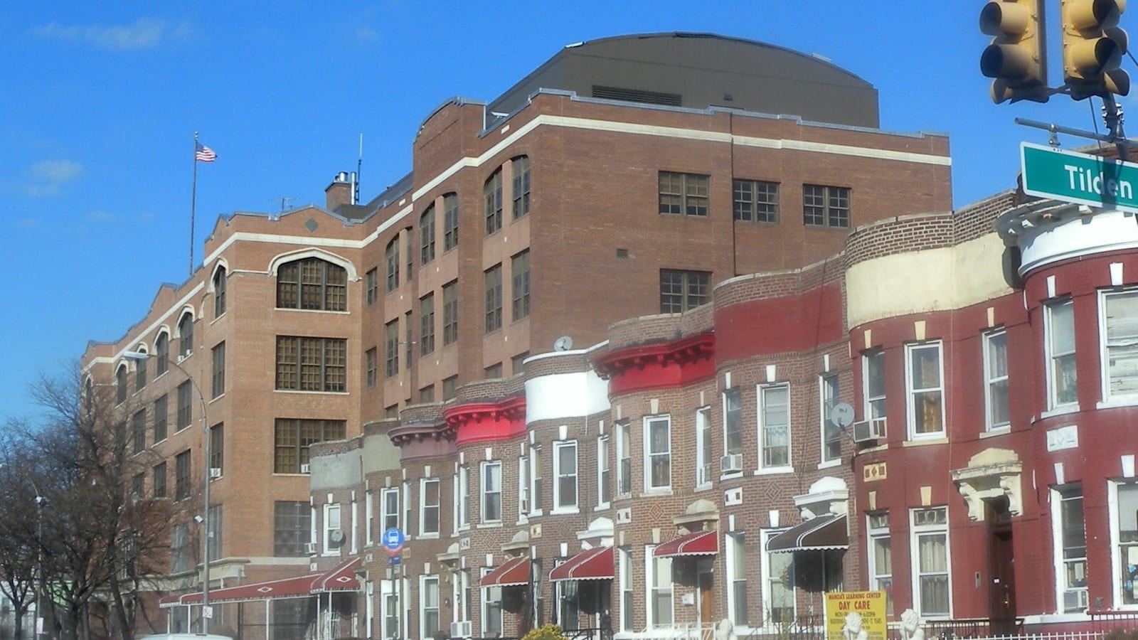 Photograph of a five-story tan brick school building with arched upper windows and a flag on its roof, behind a row of two-story brick row houses with red, white and cream bay fronts and red awnings, a traffic light and a green street sign reading Tilden at right, under a blue sky