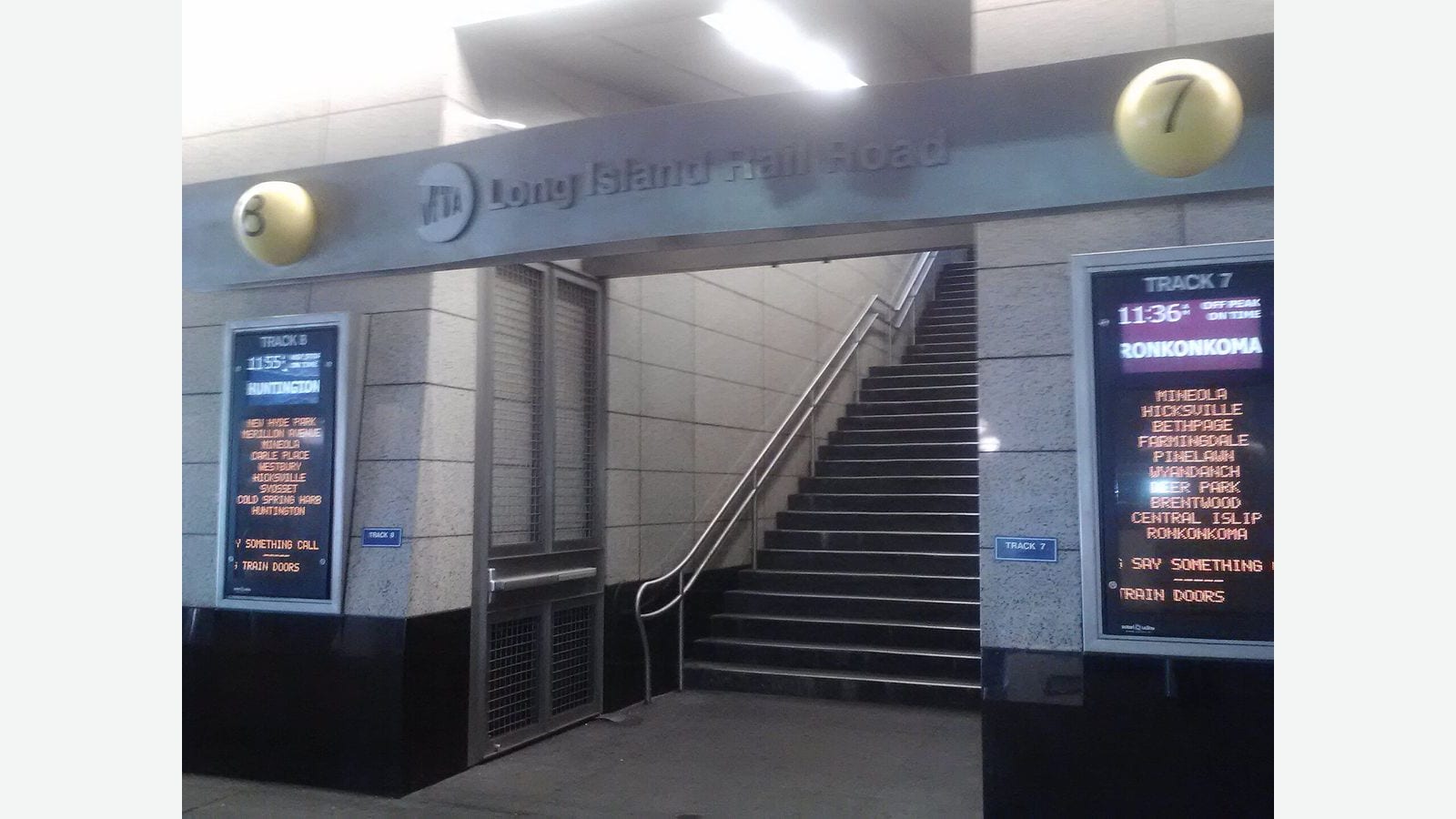Photograph of a stairway up to Long Island Rail Road tracks 7 and 8 under a grey sign reading MTA Long Island Rail Road, with yellow track-number balls and departure screens for Ronkonkoma and Huntington trains on each side