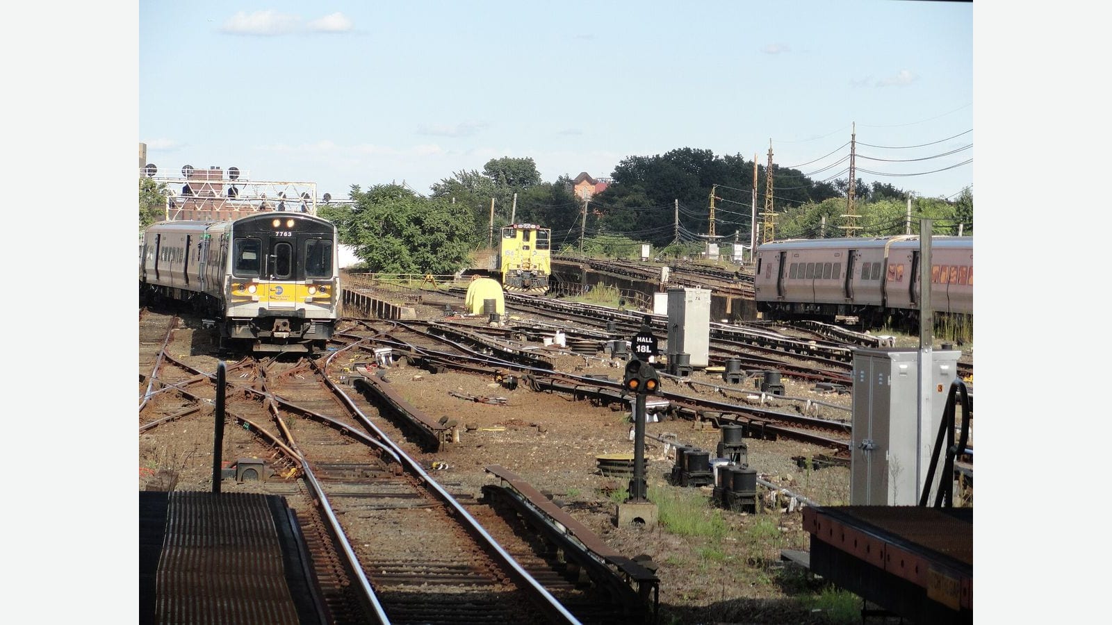Photograph of railroad tracks and switches seen from the end of a station platform, with a silver and yellow commuter train approaching on the left, a yellow work locomotive in the middle, parked passenger cars on the right, a signal marked HALL 18L and trees behind