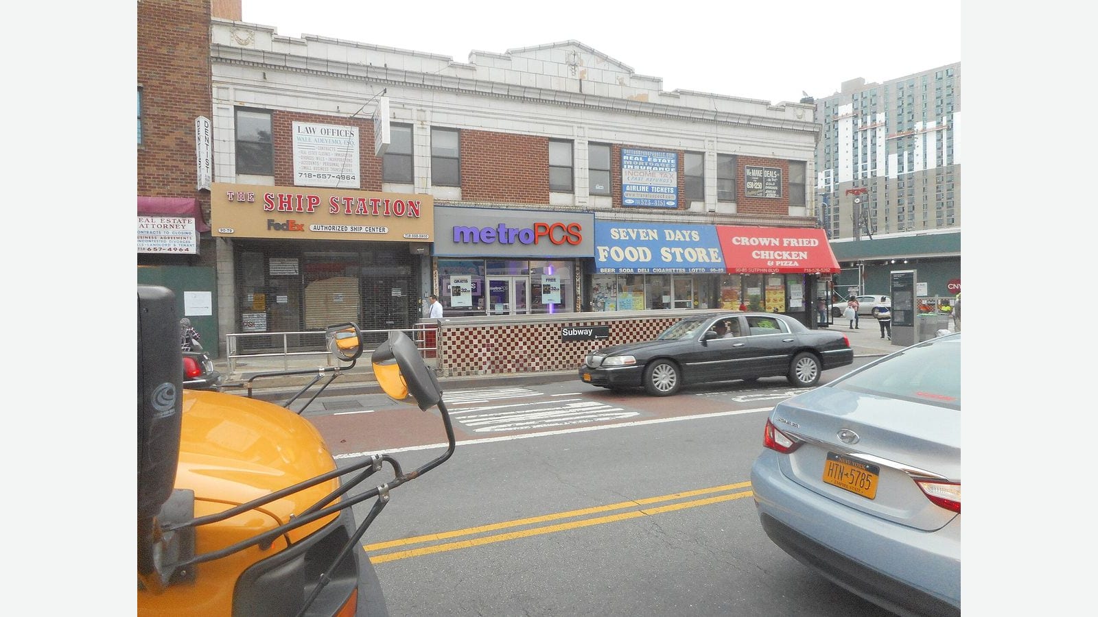 Photograph of a street-level subway entrance with a red and cream checkered tile wall and a black Subway sign on Sutphin Boulevard, in front of a two-story row of shops with signs for The Ship Station, metroPCS, Seven Days Food Store and Crown Fried Chicken, with cars on the street and the front of a yellow school bus in the lower left