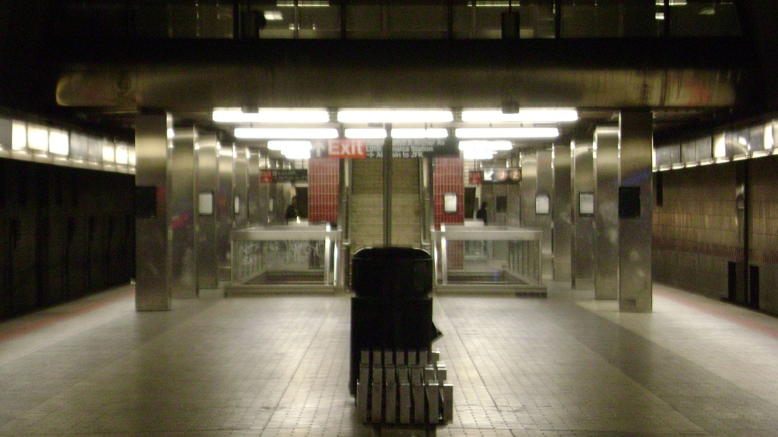 Photograph of an empty underground subway platform with steel-clad columns, bright ceiling lights, a bench and trash can in the middle, and stairs at the far end under a red Exit sign pointing to Sutphin Blvd and Archer Av, the LIRR Jamaica Station and the AirTrain to JFK