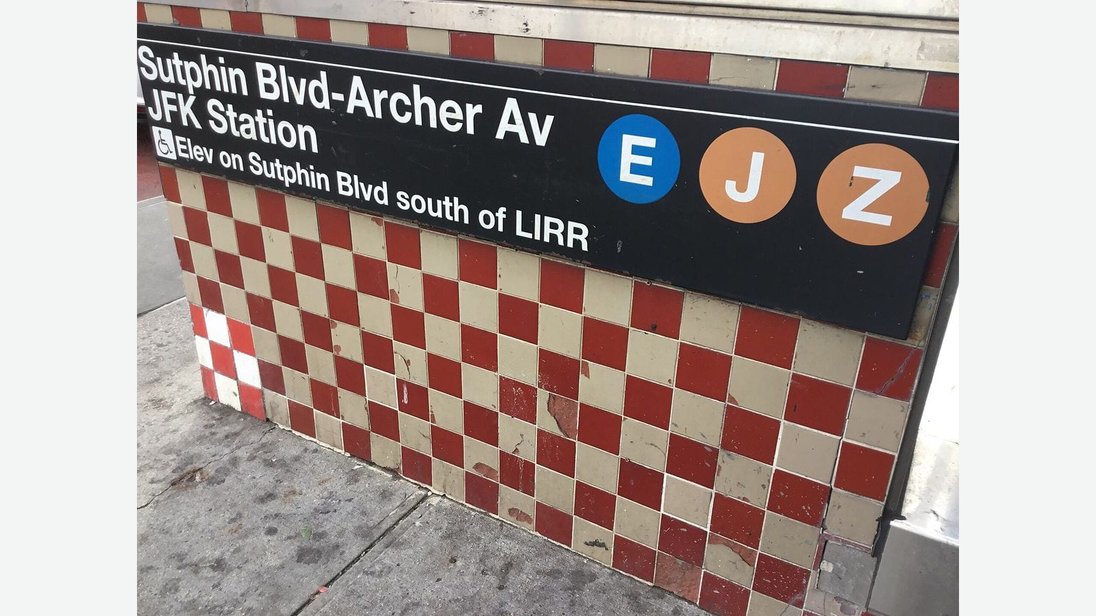 Photograph of a black station sign reading Sutphin Blvd-Archer Av JFK Station with round E, J and Z line symbols and a line reading Elev on Sutphin Blvd south of LIRR, mounted above a red and cream checkered tile wall at a sidewalk