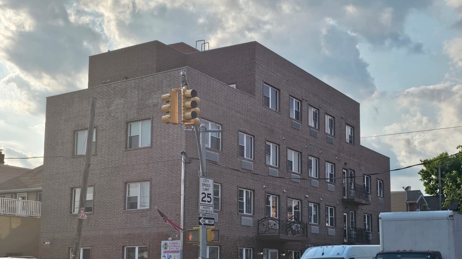 Photograph of a four-story red-brown brick apartment building on a street corner under a cloudy sky, with three full floors of white-framed windows and air-conditioner sleeves, a fourth floor set back from the front, small black iron balconies on the long side, a traffic signal, a speed-limit sign and a one-way sign on the corner pole, and the roofs of two parked vans at the bottom