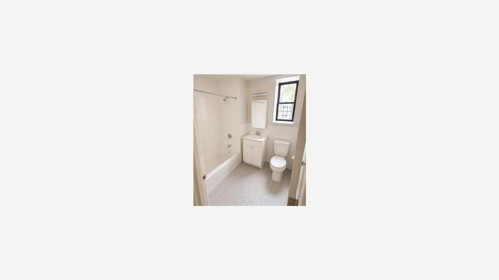 Photograph of a small white bathroom with white wall tile, a bathtub with a shower head and curtain rod, a white vanity with a mirror cabinet and light, a toilet, white hexagon floor tile and a barred window, shown small in the middle of a plain light background