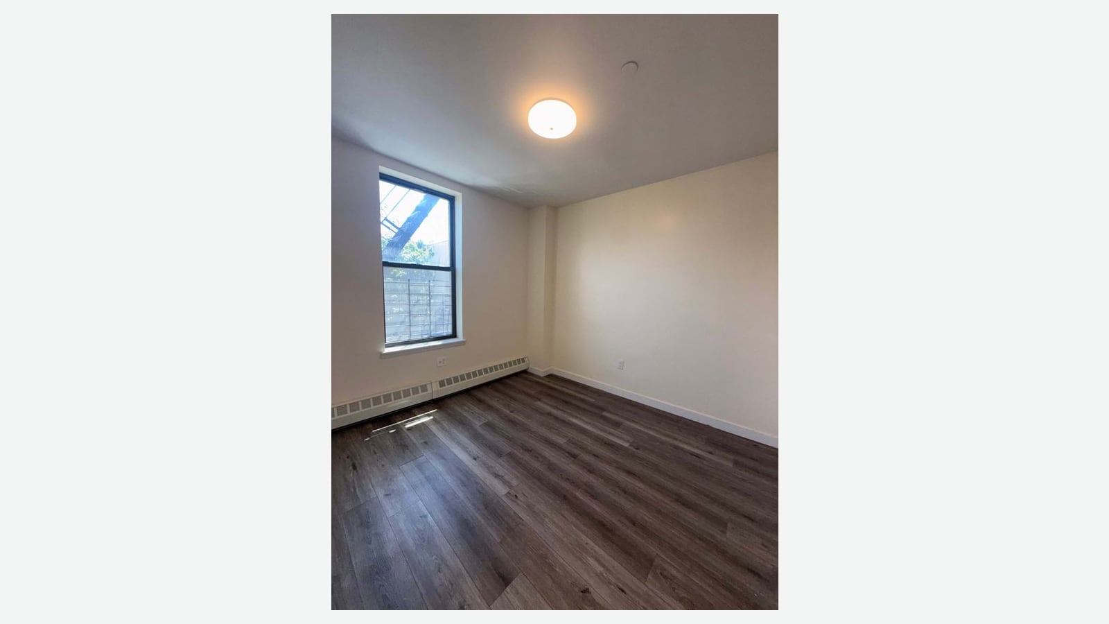 Photograph of an empty bedroom with cream walls, dark wood-look plank floors, a round ceiling light, a baseboard heater and one window with a fire escape and a tree outside, shown in the middle of a plain light background
