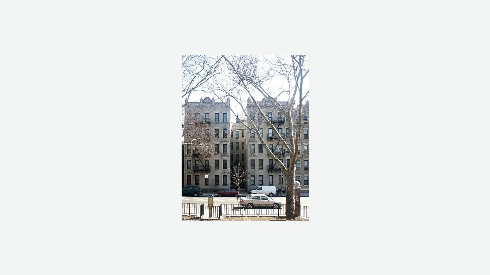 Photograph of two six-floor pale brick apartment buildings with ornate rooflines and black fire escapes, joined by a narrow recessed middle section, seen through bare winter trees from behind a low black metal fence, with parked cars and a white van along the street, shown small in the middle of a plain light background
