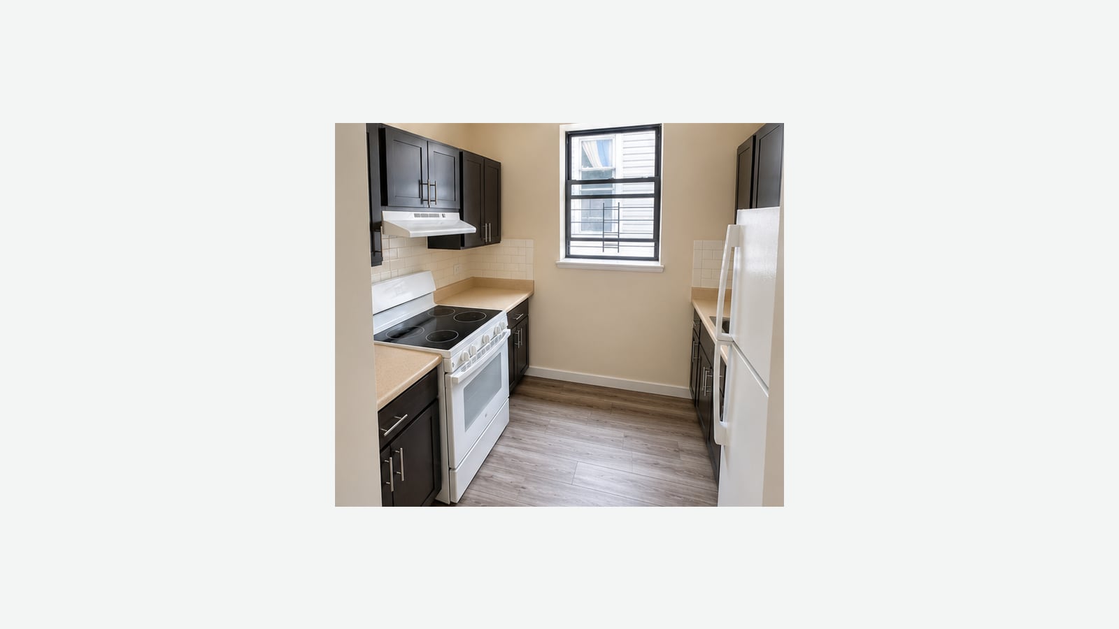 Photograph of an empty galley kitchen with dark wood cabinets, beige counters and tile backsplash, a white electric range with a smooth black cooktop under a range hood, a white refrigerator at right and a barred window at the back, shown small in the middle of a plain light background