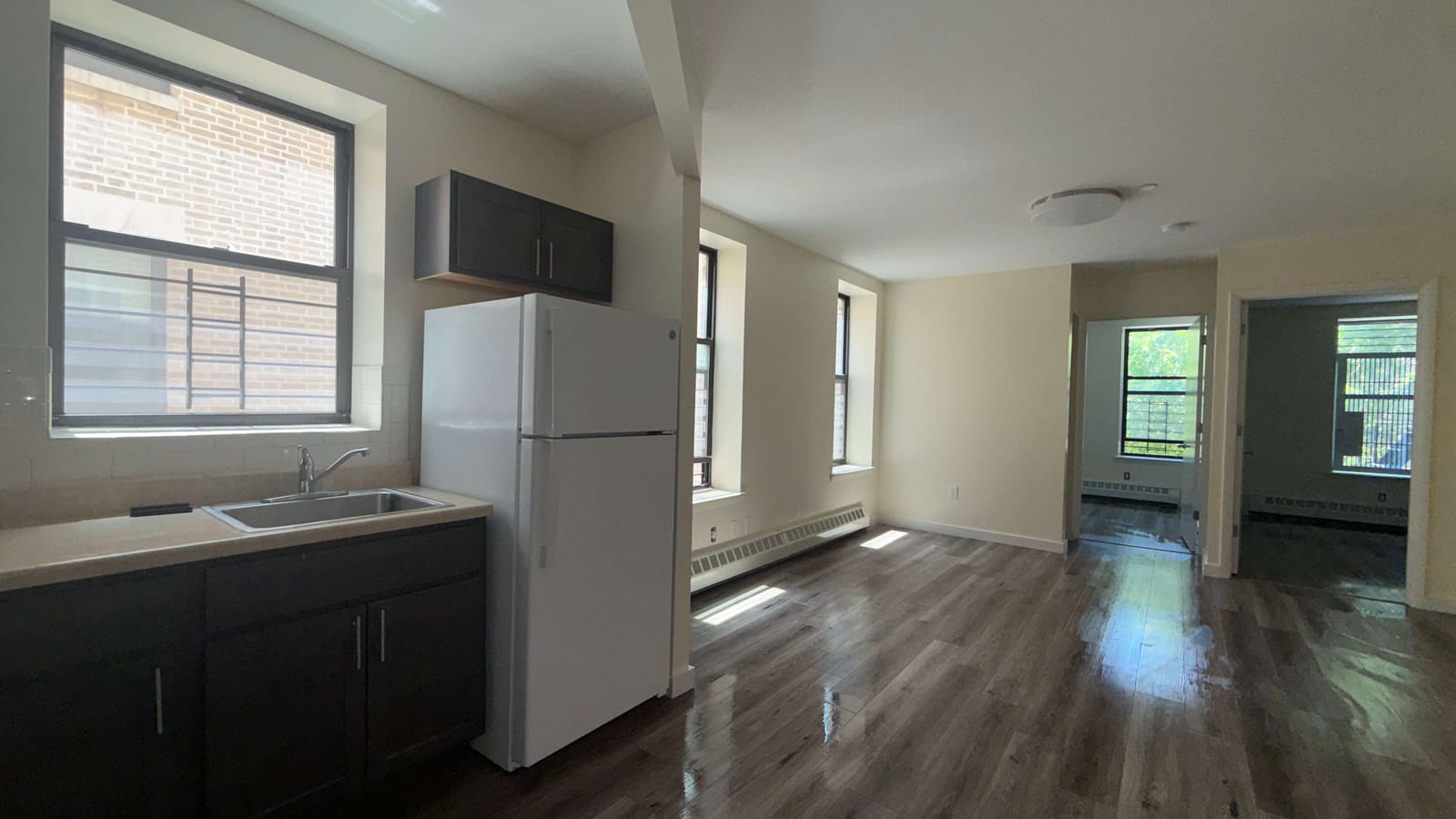 Photograph of an empty, newly finished apartment: a kitchen corner at left with dark cabinets, a light counter with a steel sink under a large barred window, a white refrigerator and a wall cabinet, opening onto a cream-colored living room with dark wood-look plank floors, a baseboard heater, two windows and two open doors to small rooms with windows