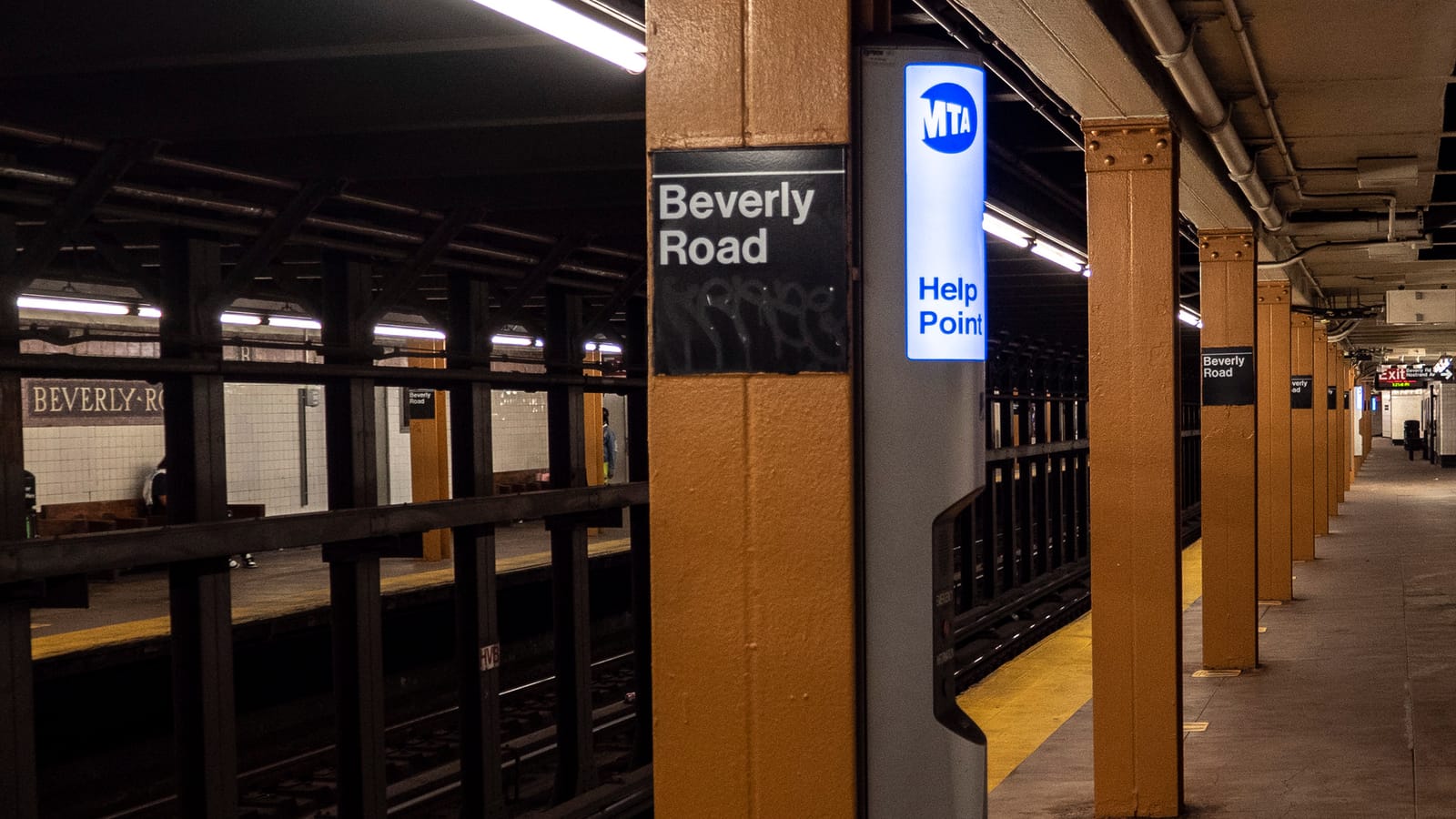Photograph of an underground subway platform with mustard-yellow steel columns, a black sign reading Beverly Road on a column, a lit blue MTA Help Point, a yellow platform edge, the opposite platform behind a railing with a tiled Beverly Road name band, and an exit sign at the far end