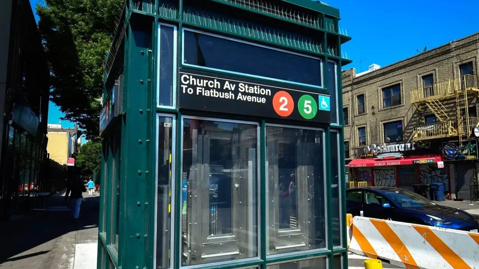 Photograph of a green glass-and-steel subway entrance kiosk on a sidewalk under a blue sky, with a sign reading Church Av Station To Flatbush Avenue, round 2 and 5 route symbols and a wheelchair symbol, an orange and white barrier, a parked blue car and a three-story tan brick building with a restaurant awning behind it