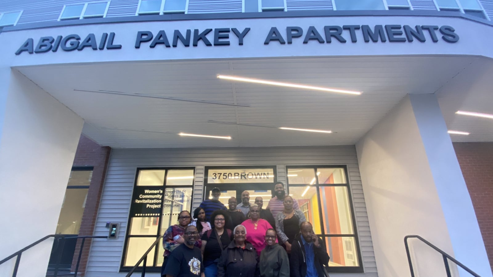 Building entrance under a white canopy with raised letters reading Abigail Pankey Apartments, glass doors marked 3750 Brown, and a group of people standing on the steps