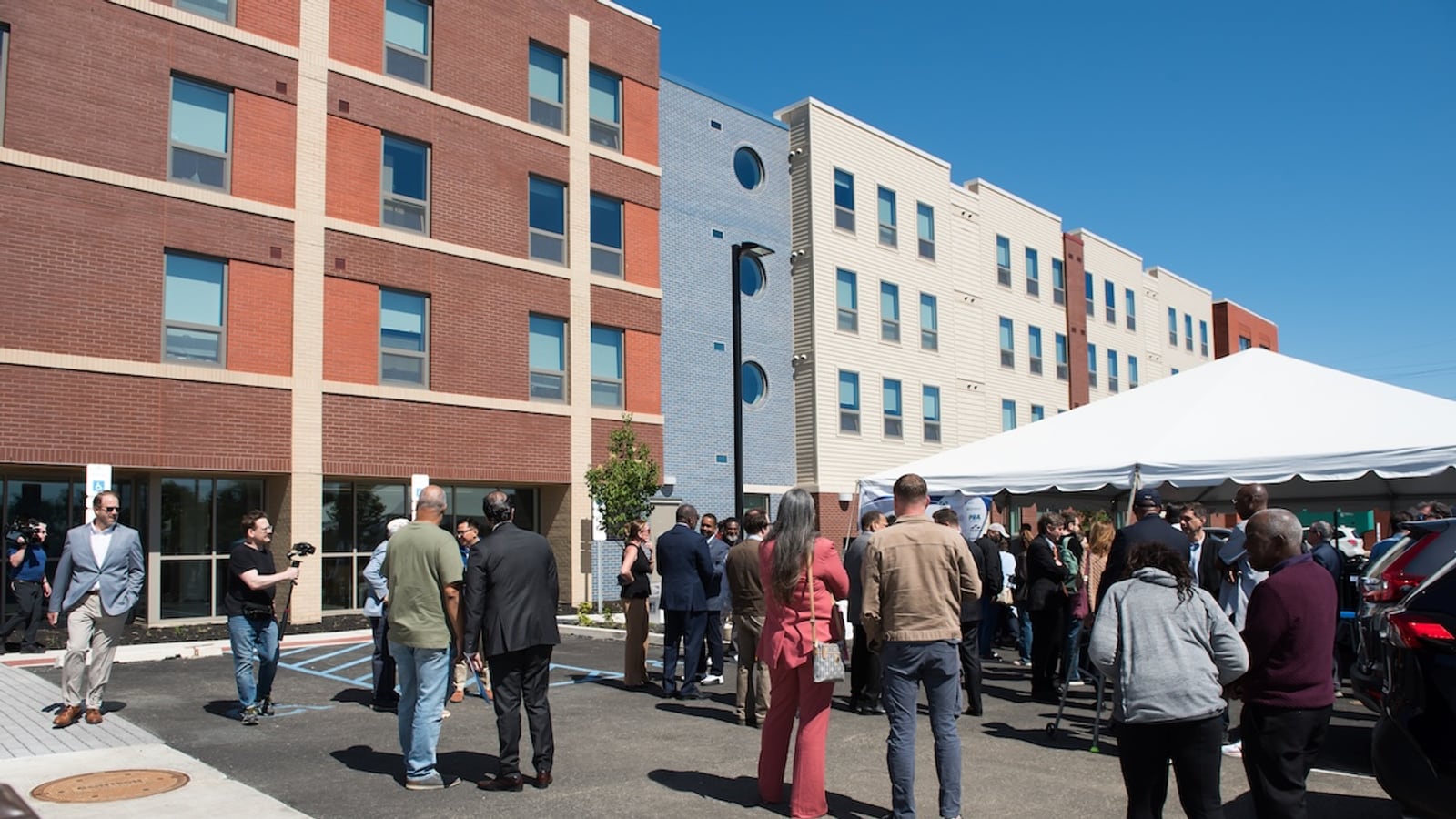 People standing in a parking lot in front of a four-story brick and sided apartment building, with a white tent at the right