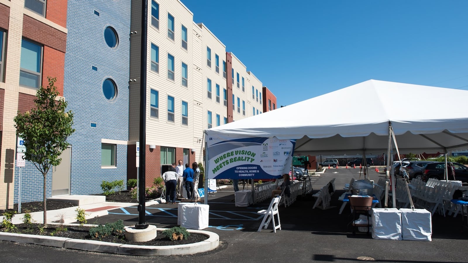 Side of a four-story building with a blue brick section and round windows beside pale siding, with a white event tent set up in the parking lot
