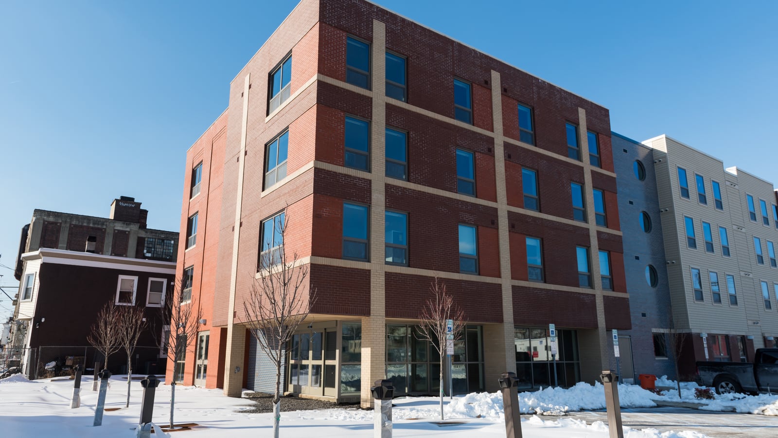 Four-story apartment building in red brick with tan vertical bands and a lighter sided wing with round windows, seen from a snow-covered corner under a blue sky
