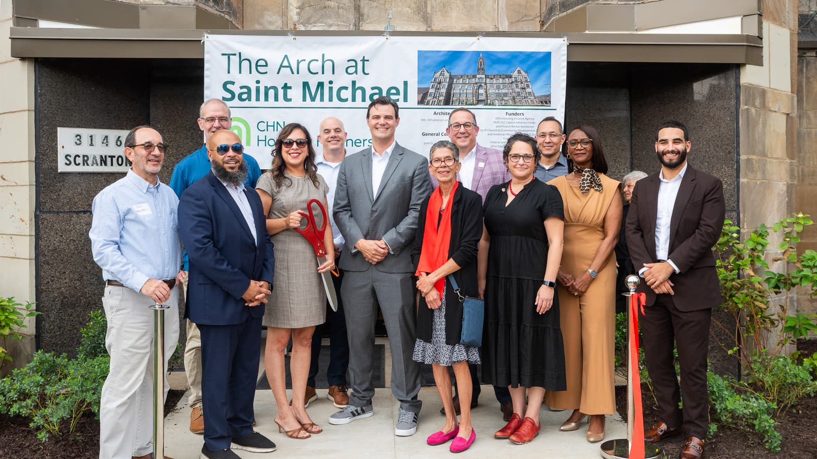 Twelve people standing at a building entrance marked 3146 Scranton under a banner reading The Arch at Saint Michael, one holding large red scissors beside a red ribbon