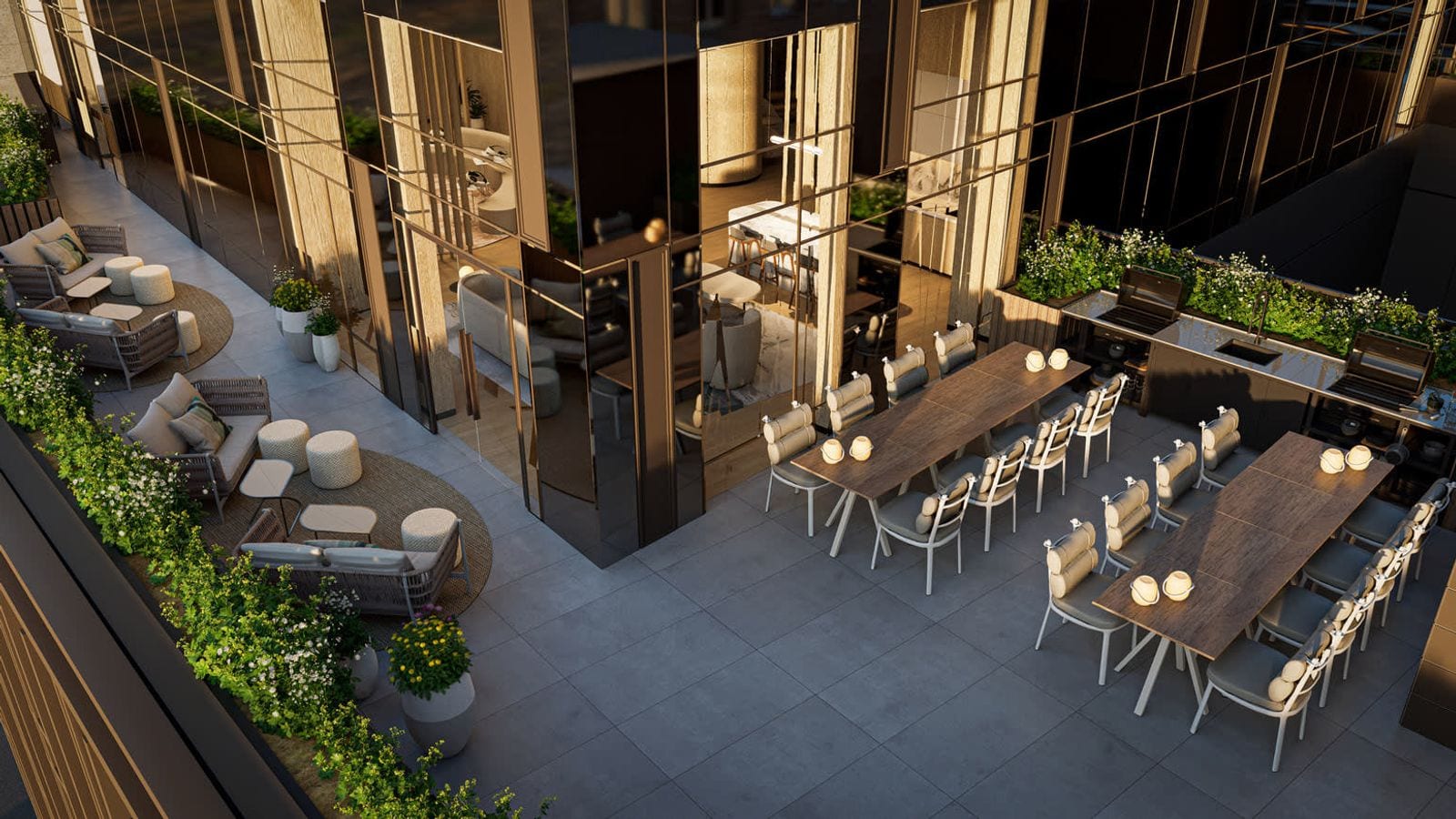 Rendering looking down on an outdoor terrace at dusk: two long dining tables with white chairs, a counter with two grills and a sink in front of a planted hedge, sofas and round stools along a planted edge, and dark glass walls with a lit lounge behind them
