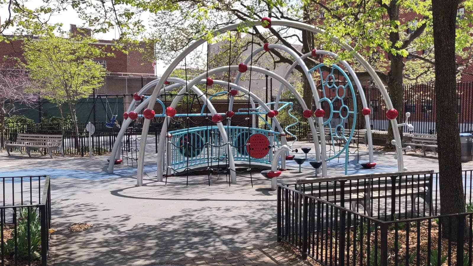 Photograph of a playground with large silver arches dotted with red balls, blue climbing panels and rope nets over a gray safety surface, wooden benches, a low black iron fence and trees in spring leaf, with brick buildings behind