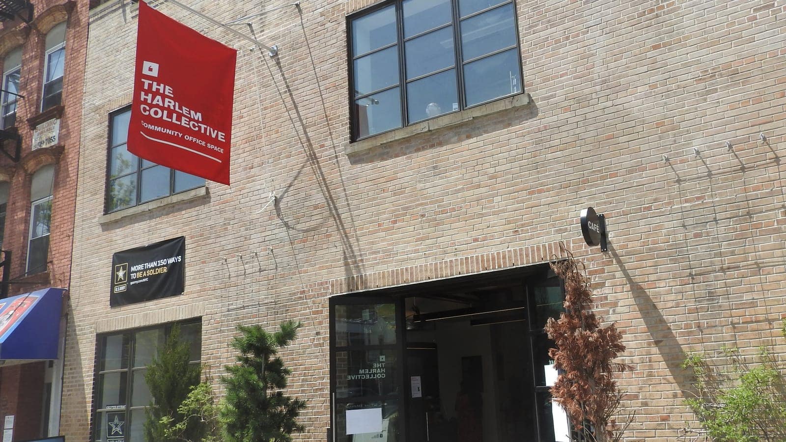 Photograph of a tan brick building with large black-framed windows, a red hanging banner reading The Harlem Collective Community Office Space, a round sign reading Cafe over an open glass entrance, small evergreen and dried plants in front and a red brick row building at left